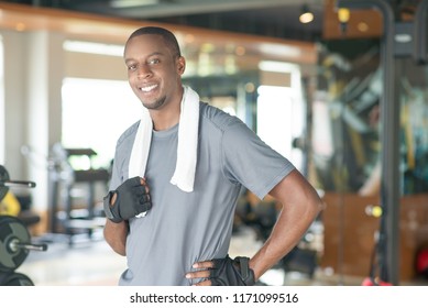 Smiling Sporty Black Man Wearing Towel Around Neck And Looking At Camera. Young Guy Wearing T-shirt And Relaxing In Gym. Workout And Break Concept. Front View.