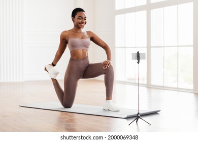 Smiling Sporty Black Woman Training At Home In Front Of Smartphone On Tripod, Beautiful Female Fitness Blogger Shooting Online Video Fitness Tutorials, Showing Legs Quad Stretching Exercises At Camera