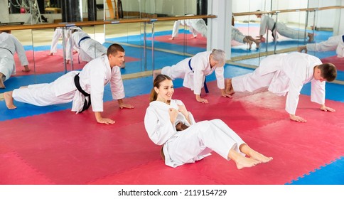 Smiling sporty young woman in white kimono warming up before group martial arts training in gym, doing abdominal crunch..