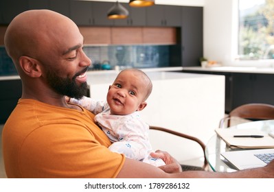 Smiling Stay At Home African American Father Cuddling Baby Daughter Whilst Working On Laptop At Home