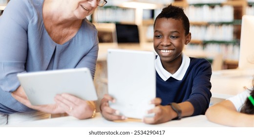 Smiling student boy uses a tablet in a library, guided by woman teacher. Diverse teacher and student hold tablets, engaging, learning activity together in the library. Teacher teaching boy in class - Powered by Shutterstock