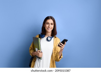 Smiling student girl is holding mobile phone in hand, looks at camera, learning online through application, holding books, wearing yellow shirt, white t-shirt, black bag and headphones over neck - Shutterstock ID 2095015387