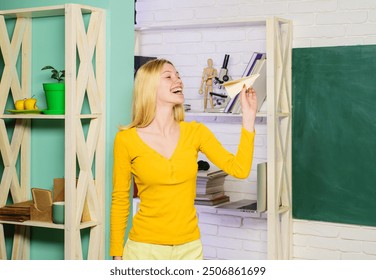 Smiling student girl playing with paper airplane. Happy girl throws origami plane. Young woman with paper airplane in classroom. Schoolgirl launches paper aeroplane near chalkboard. Dreams of travel. - Powered by Shutterstock