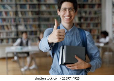 Smiling student guy in glasses holds books and textbooks showing thumbs up gesture at camera posing in campus library, close up finger gesture. Higher institution learner, education, knowledge concept