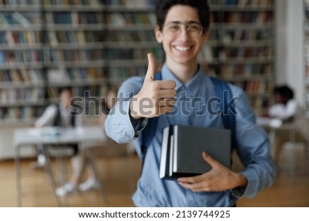 Smiling student guy in glasses holds books and textbooks showing thumbs up gesture at camera posing in campus library, close up finger gesture. Higher institution learner, education, knowledge concept