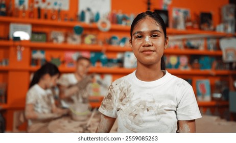 Smiling student looking at camera while standing at workshop while diverse student having pottery class together. Happy caucasian girl smile while wear dirty shirt in art lesson. Edification. - Powered by Shutterstock