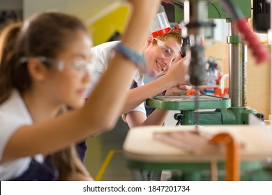 A smiling student with safety glasses bends down to have a closer look while working on a drill machine