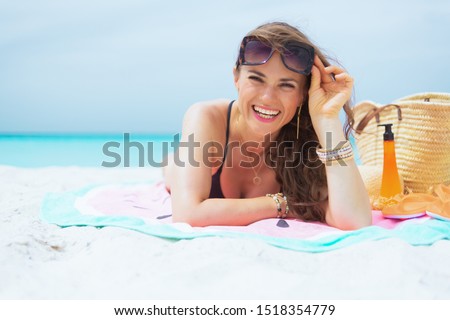 smiling stylish 40 year old woman with long curly hair in elegant black bathing suit on a white beach sun tanning while laying on a round towel.