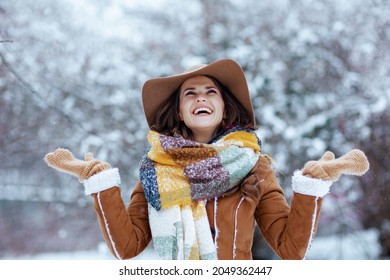 smiling stylish 40 years old woman in brown hat and scarf with mittens in sheepskin coat catching snow outdoors in the city park in winter.