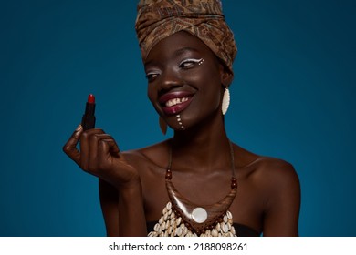 Smiling stylish black girl holding lipstick. Attractive young slim woman wearing traditional african outfit and accessories. Female beauty. Isolated on blue background. Studio shoot. Copy space