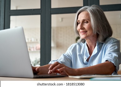 Smiling Stylish Mature Middle Aged Business Woman Using Laptop Computer Sitting At Workplace Desk. Happy Senior Older Lady, 60s Grey-haired Businesswoman Working Typing On Pc At Home Or From Office.