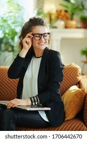 Smiling Stylish Middle Age Woman In White Blouse And Black Jacket With Closed Laptop Sitting On Couch At Modern Home In Sunny Day.