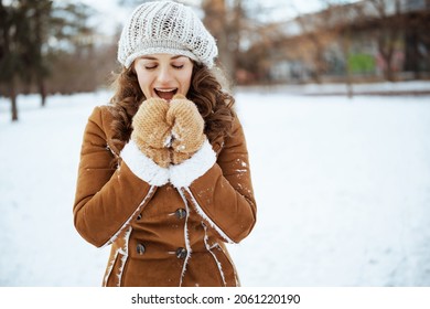 smiling stylish middle aged woman with mittens in a knitted hat and sheepskin coat warming hands with breath outside in the city park in winter.