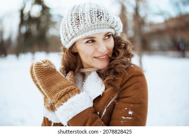 Smiling Stylish Middle Aged Woman With Mittens In A Knitted Hat And Sheepskin Coat Outside In The City Park In Winter.