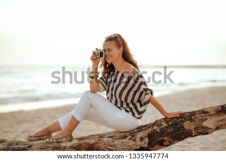 smiling stylish woman in white pants and striped blouse taking photos with retro photo camera while sitting on a wooden snag on the seashore at sunset.