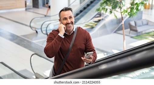 Smiling successful business man going up in escalator talking on smart phone while using wireless earbuds at airport. Happy entrepreneur talking over mobile phone while going to work.