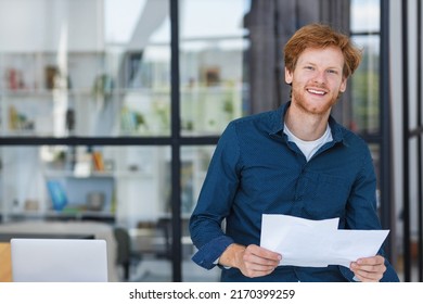 Smiling successful businessman stands near desk smiling and looking at the camera. Young positive male confident entrepreneur or small business owner in a modern office or coworking space