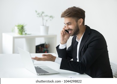 Smiling successful businessman in suit talking on phone using laptop sitting at office workplace, young entrepreneur making answering call consulting client speaking by cell, holding mobile interview