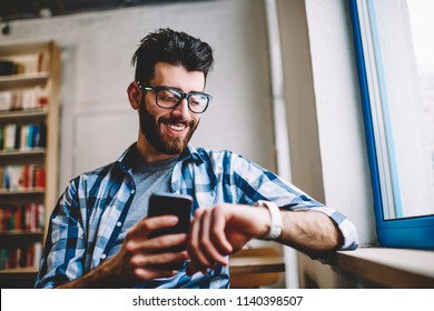 Smiling successful hipster guy in eyewear checking time on wearable smartwatch while waiting for meeting at university campus, happy man holding mobile phone in hand and reading message on wristwatch