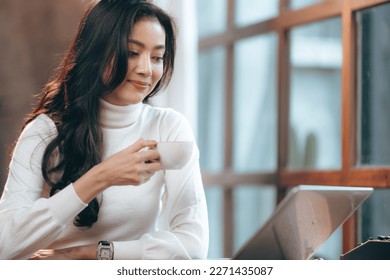 Smiling and successful young female Asian entrepreneur working on laptop while taking a break by sipping coffee and tea and looking away while thinking about new strategy and relaxing - Powered by Shutterstock