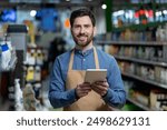 Smiling supermarket worker using digital tablet for inventory management in store aisle. Employee wearing apron holding tablet, performing inventory management tasks. Modern retail technology