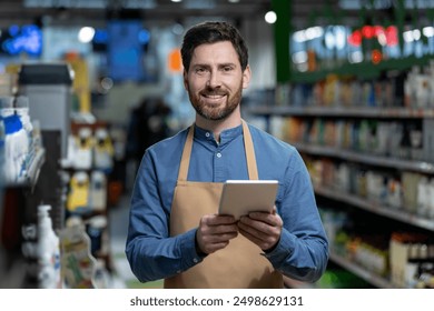 Smiling supermarket worker using digital tablet for inventory management in store aisle. Employee wearing apron holding tablet, performing inventory management tasks. Modern retail technology - Powered by Shutterstock