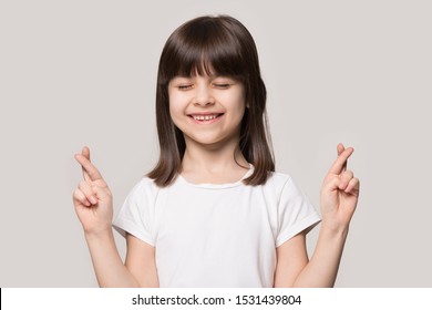 Smiling Superstitious Little Girl Isolated On Grey Studio Background Cross Fingers Close Eyes Make Wish, Happy Cute Small Child Believer Make Gesture Hope Believe Ask For Good Luck, Faith Concept