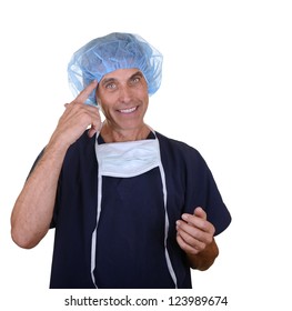 Smiling Surgeon Wearing Navy Blue Scrubs, Hair Cover, And Mask Hanging Down Pointing At Himself