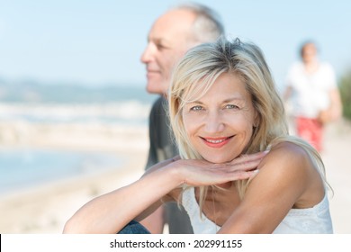 Smiling tanned mature woman at the beach with her husband turning to smile at the camera as they enjoy a healthy active lifestyle
