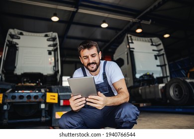 Smiling Tattooed Bearded Blue Collar Worker In Overalls Using Tablet To Check On Delivery While Crouching In Garage Of Import And Export Firm. In Background Are Trucks.