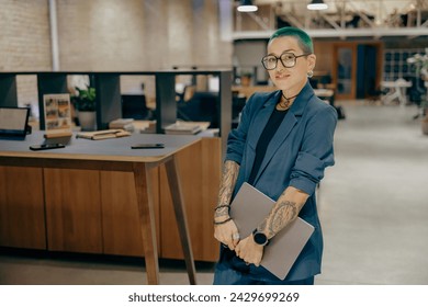 Smiling tattooed business woman with green short hair holding laptop standing in modern office - Powered by Shutterstock