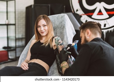 Smiling tattooed girl looking in camera while professional tattooer doing tattoo on hand using tattoo machine in studio