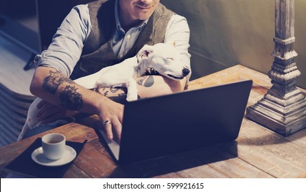 Smiling Tattooed Man In Eyeglasses Working At Home On Laptop While Sitting At The Wooden Table With Cute Dog Sleeping On His Hands.Adult Guy Using Modern Computer For Surfing Web.Horizontal