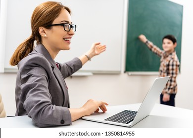 Smiling teacher in glasses with laptop pointing with hand at blackboard