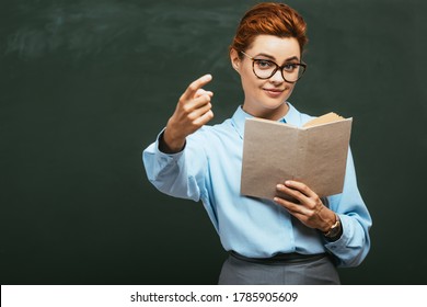 Smiling Teacher Holding Open Book And Showing Come Here Gesture While Standing Near Chalkboard