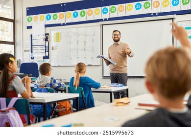 Smiling teacher holding textbook pointing towards boy raising hand to answer. Happy teacher aiming at his elementary students while wanting to hear the answer to his question. Elementary classroom. - Powered by Shutterstock