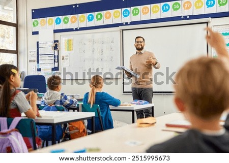 Smiling teacher holding textbook pointing towards boy raising hand to answer. Happy teacher aiming at his elementary students while wanting to hear the answer to his question. Elementary classroom.