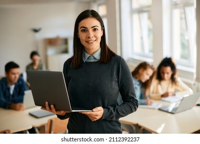 Smiling teacher using laptop during computer class at high school and looking at camera. Her students are learning in the background.  - Shutterstock ID 2112392237
