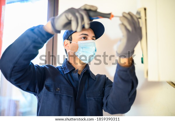 Smiling technician repairing an\
hot-water heater wearing a mask due to coronavirus\
pandemic