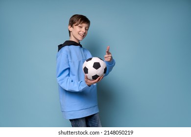 Smiling Teen Boy In Sports Blue Clothes In A Sweatshirt Holding Soccer Ball Posing In Studio On A Colored Background. Caucasian Teenager Fan Of Playing Football