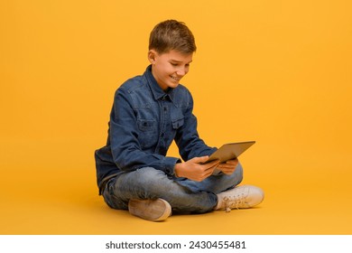 Smiling teen boy using digital tablet while sitting cross-legged on floor in studio, happy teenage male kid engaged in digital learning or entertainment, posing against yellow background - Powered by Shutterstock