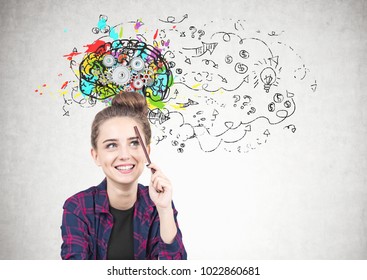 Smiling teen girl in checkered shirt is sitting and pencil near her forehead   thinking  She is looking at colorful brain sketch and cogs it   business idea sketch 