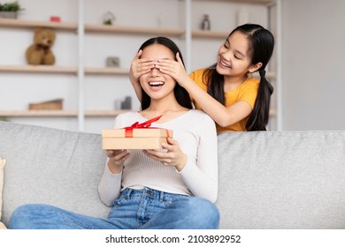 Smiling Teen Girl Congratulates Surprised Young Asian Lady, Gives Box With Present, Closes Eyes To Mom Sit In Room Interior. Happy Birthday, Family Relationships, Congratulations At Home Due Covid-19