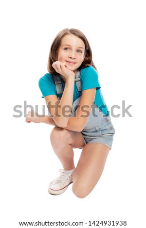 Smiling teen girl in denim overalls squats. Isolated on a white background. Vertical.
