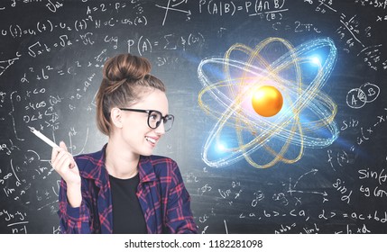 Smiling teen girl looking at gold and blue atom model over blackboard with formula background. Concept of science, chemistry and physics education. Toned image double exposure