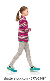 Smiling Teen Girl In Striped Fleece Jacket, Jeans And Sneakers Walking And Looking Away. Side View. Full Length Studio Shot Isolated On White.