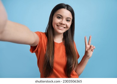 Smiling teen girl taking selfie showing peace sign with two fingers looking at camera on mobile phone, isolated over blue color background - Powered by Shutterstock