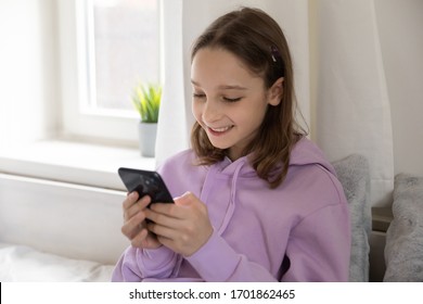 Smiling teen girl using mobile phone apps at home. Happy schoolgirl holding smartphone texting message, chatting with friends in social media, playing game or watching stories looking at cellphone.