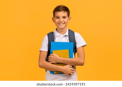 Smiling teen schoolboy with backpack holding textbooks and smiling at camera, happy male kid dressed in white polo standing against yellow studio background, portraying enthusiasm for education - Powered by Shutterstock