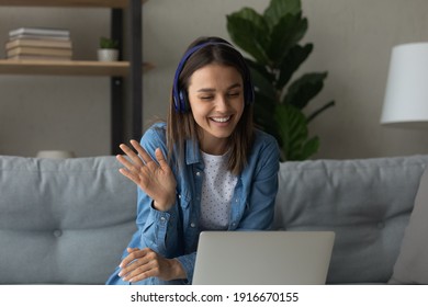 Smiling Teenage Female Pupil Sit On Sofa In Headphones Wave Hand To Laptop Screen Greet Teacher Tutor At Lesson Consultation. Happy Young Woman Say Hi Hello To Friend Speaking From Home By Video Call